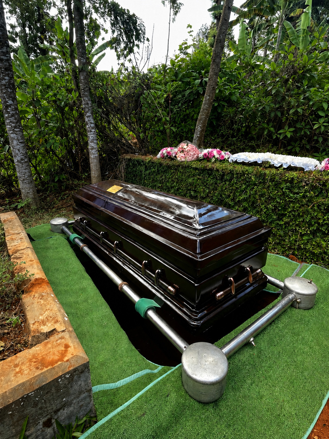 Casket resting on lowering equipment at a graveside prepared with green artificial grass carpeting and floral tributes